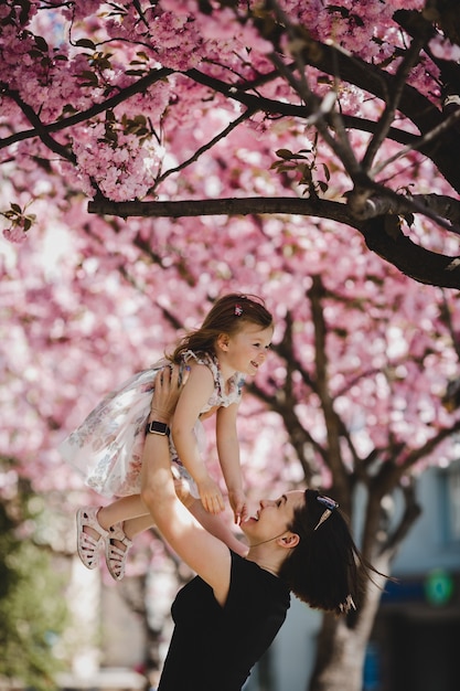 Beautiful young mom holds lovely little daughter standing under blooming pink tree