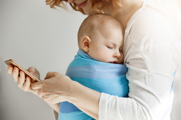 Beautiful young mother reads articles about maternity life on phone while little lovely son sleeping on her chest in blue baby sling.