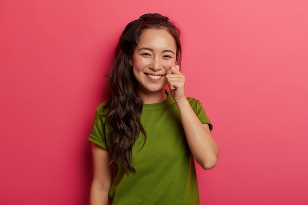 Beautiful young natural brunette girl touches cheek gently, has healthy skin, shapes mini heart with fingers isolated over vivid pink background.