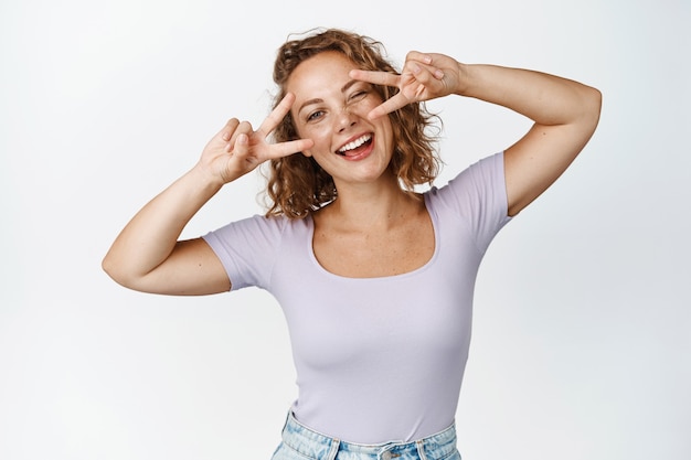 Bella giovane donna naturale con capelli biondi ricci, strizzando l'occhio, mostrando la pace del v-segno vicino agli occhi, sorridendo felice su bianco.