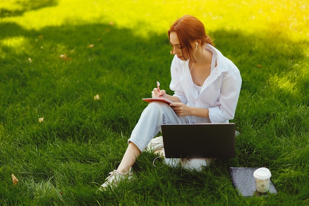 有線で勉強や仕事のためにラップトップコンピューターを使用して屋外の公園で美しい若いかなり赤毛の女性...