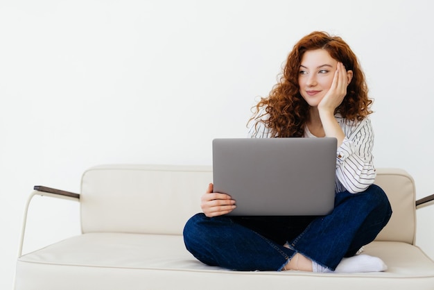 Красивая молодая рыжеволосая женщина сидит дома на диване и печатает на ноутбуке