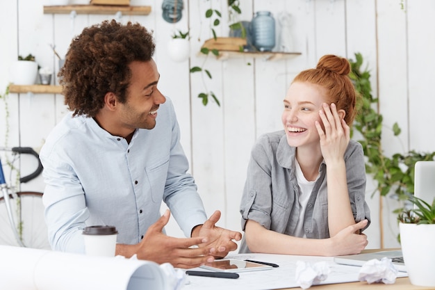 Beautiful young redhead woman designer laughing at jokes of her cheerful dark-skinned male colleague