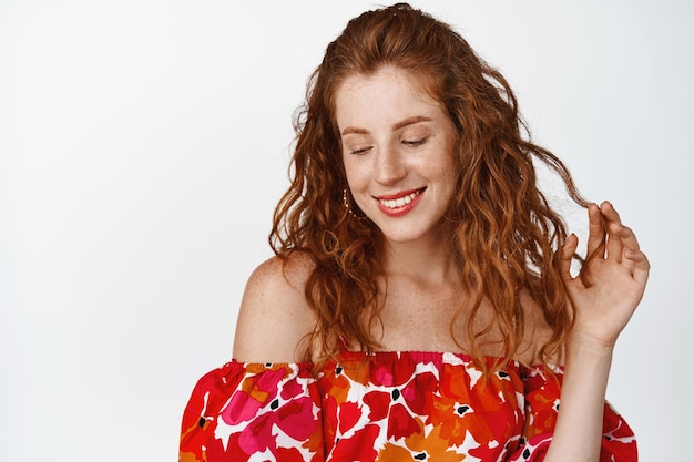 Красивая молодая рыжая женщина, играющая со своими вьющимися волосами, смотрит вниз с кокетливой кокетливой улыбкой, стоящей на белом фоне