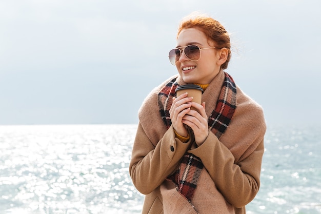 Красивая молодая рыжеволосая женщина в пальто гуляет на пляже, пьет кофе на вынос
