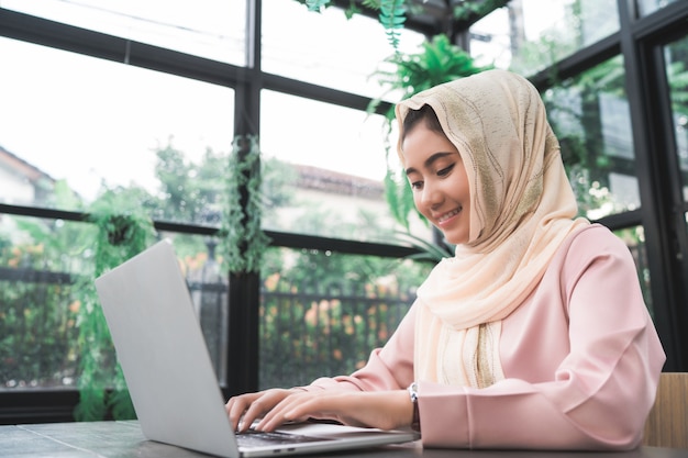 Красивая молодая женщина улыбается азиатских мусульман, работающих на ноутбуке, сидя в гостиной дома