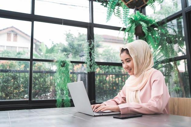 Beautiful young smiling asian muslim woman working on laptop sitting in living room at home