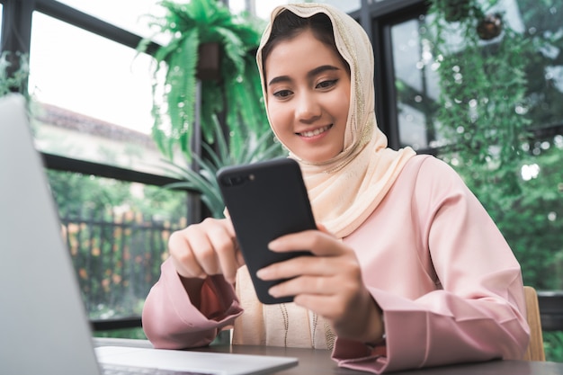Beautiful young smiling asian muslim woman working on phone sitting in living room at home