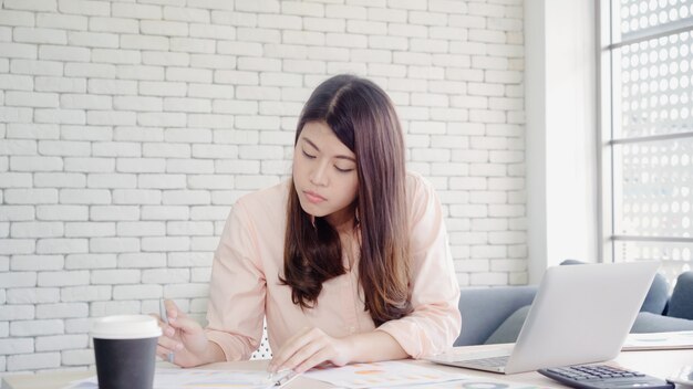 Beautiful young smiling asian woman working laptop on desk in living room at home. 