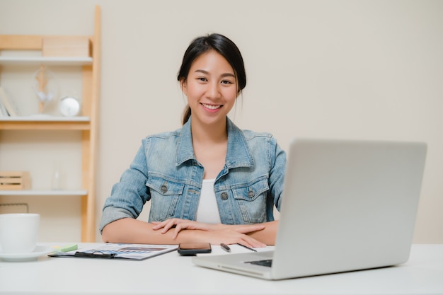 Beautiful young smiling asian woman working laptop on desk in living room at home. 