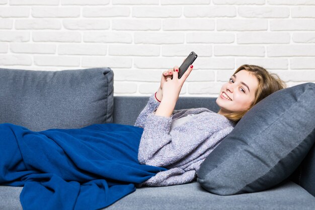 Beautiful young smiling brunette woman lying in white bed and using a phone in her bedroom.