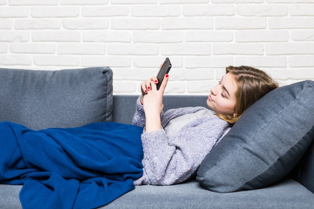 Free photo beautiful young smiling brunette woman lying in white bed and using a phone in her bedroom.