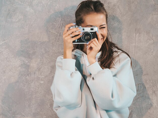 아름 다운 젊은 미소 사진 여자 그녀의 레트로 카메라를 사용 하여 사진을 찍고. 여자 사진 만들기. 캐주얼 한 여름 까마귀를 입은 모델. 회색 벽 근처 스튜디오에서 포즈