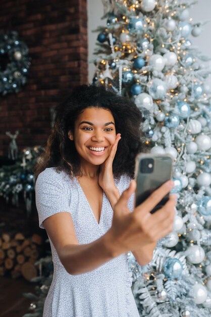 バックグラウンドでクリスマスツリーの近くの美しい若い笑顔の女性は、クリスマスの装飾の部屋で休日と新年のビデオメッセージまたはSelfieコンセプトを作成します。