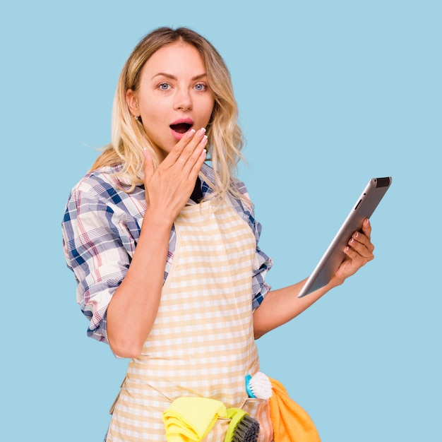 Beautiful young surprised woman wearing apron holding digital tablet against blue backdrop