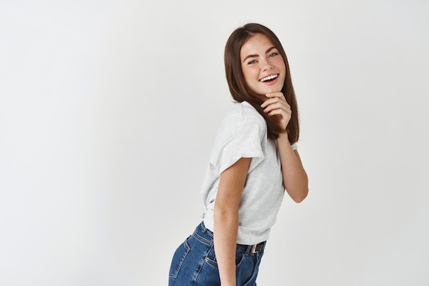 Красивая молодая татуированная женщина в белой футболке позирует на фоне студии, улыбаясь и трогая идеальное лицо без пятен на белом фоне