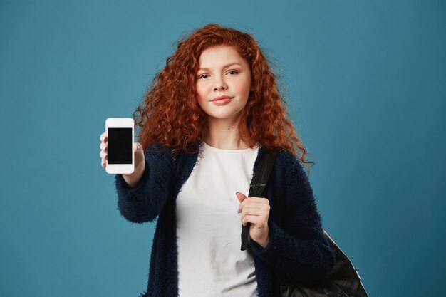 Beautiful young teen red-haired woman with freckles showing cell phone, holding backpack with hand, having happy and confident look.