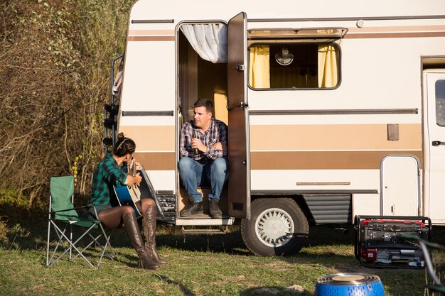 Beautiful young wife singing a song on her guitar for her husband in front of their reto camper in the mountains.