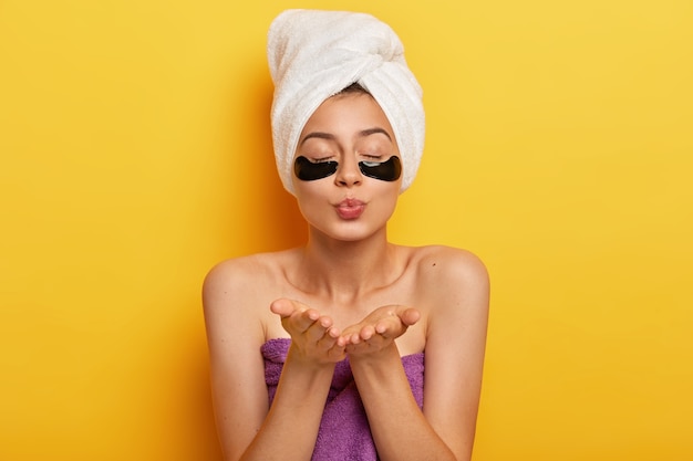 Beautiful young woman applies collagen patches for removing wrinkles, sends air kiss, keeps eyes shut, wears white towel on head