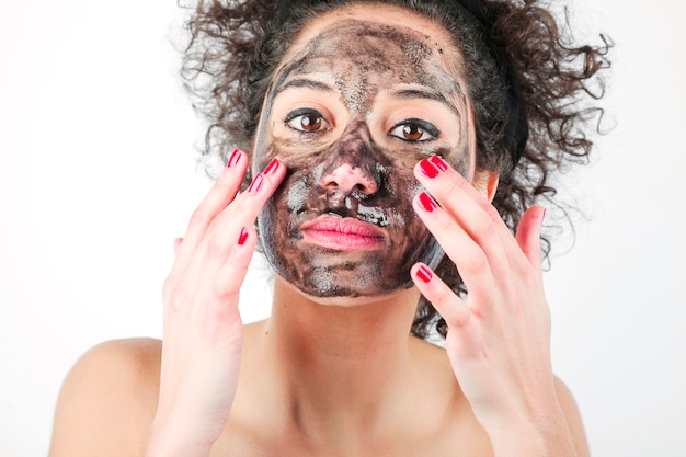 Beautiful young woman applying black face mask with her fingers against white background