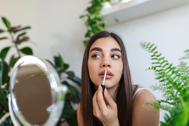 Beautiful young woman applying lip gloss with applicator in front of a mirror at home