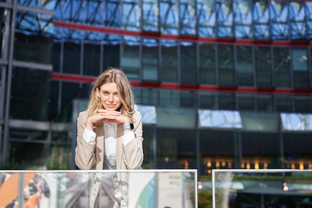 Beautiful young woman in beige suit standing near office buildings in city center smiling and lookin