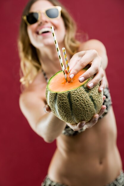 Beautiful young woman in bikini drinking melon juice. Isolated on red.