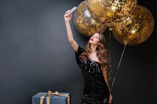 Beautiful young woman in black luxury dress, red lips, long curly brunette hair taking selfie portrait with big balloons full with golden tinsels. Party time, true emotions.