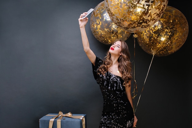 Beautiful young woman in black luxury dress, red lips, long curly brunette hair taking selfie portrait with big balloons full with golden tinsels. Party time, true emotions.