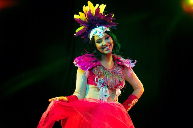 Beautiful young woman in carnival mask and stylish masquerade costume with feathers in colorful lights and glow on black wall