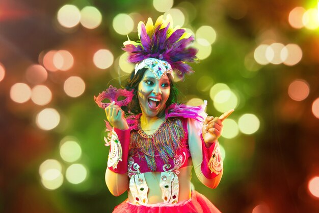 Beautiful young woman in carnival mask and stylish masquerade costume with feathers and sparklers in colorful bokeh on black background