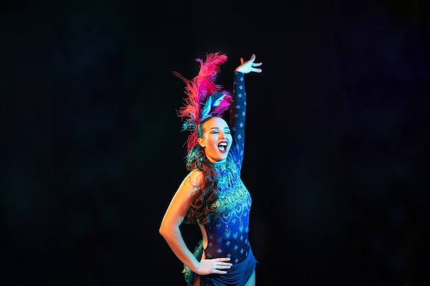 Beautiful young woman in carnival, stylish masquerade costume with feathers on black background in neon light.