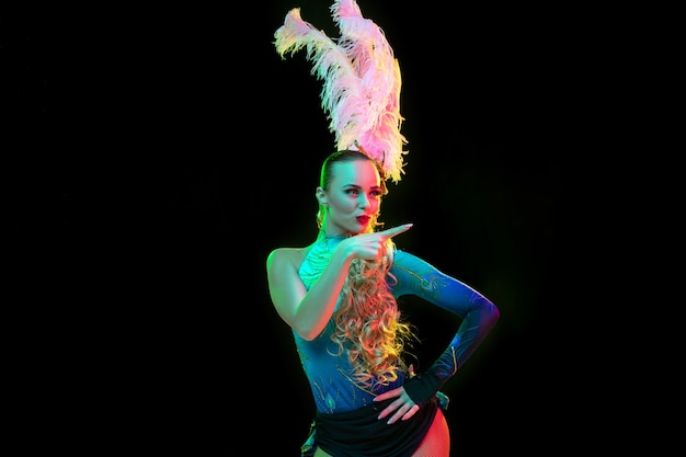 Beautiful young woman in carnival, stylish masquerade costume with feathers on black background in neon light.