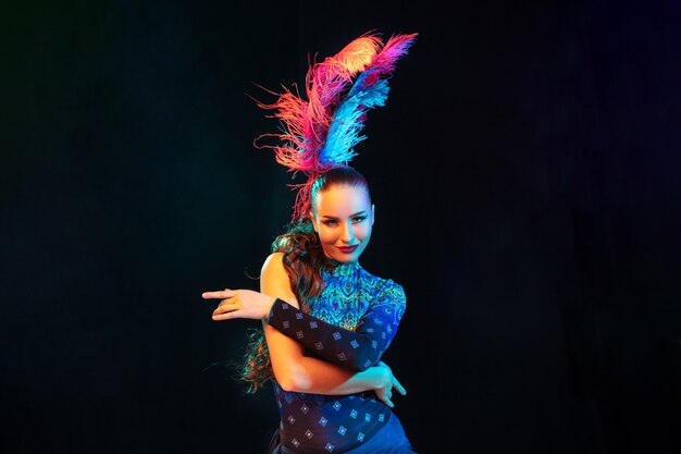 Beautiful young woman in carnival, stylish masquerade costume with feathers on black wall in neon light