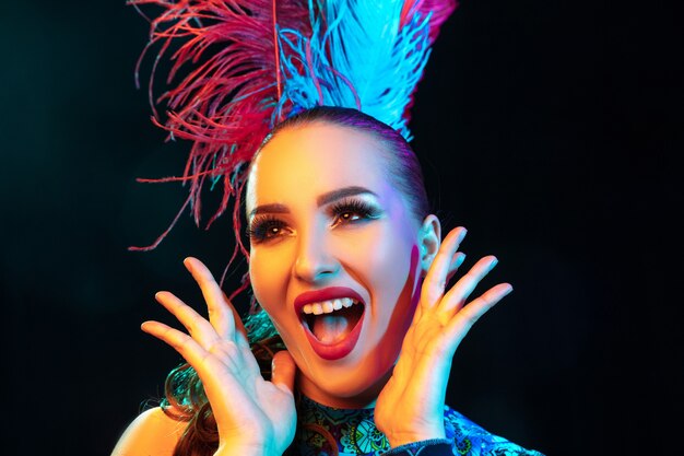 Beautiful young woman in carnival, stylish masquerade costume with feathers on black wall in neon light