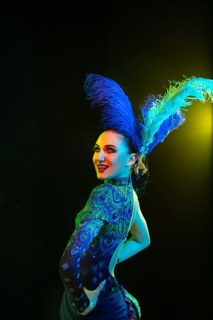 Beautiful young woman in carnival, stylish masquerade costume with feathers on black wall in neon light
