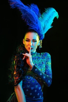 Bella giovane donna in carnevale, elegante costume in maschera con piume sul muro nero in luce al neon
