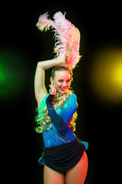 Beautiful young woman in carnival, stylish masquerade costume with feathers on black wall in neon light