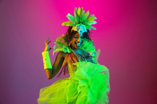 Beautiful young woman in carnival, stylish masquerade costume with feathers dancing on gradient background in neon light.