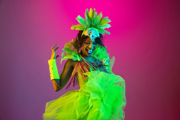 Beautiful young woman in carnival, stylish masquerade costume with feathers dancing on gradient background in neon light.