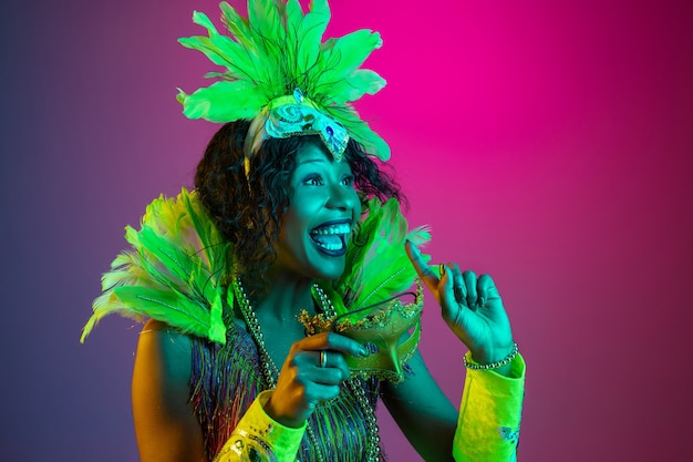 カーニバルの美しい若い女性、ネオンのグラデーションの背景に羽が踊るスタイリッシュな仮面舞踏会の衣装。