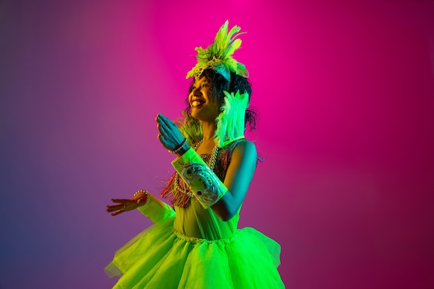 カーニバルの美しい若い女性、ネオンのグラデーションの壁に羽が踊るスタイリッシュな仮面舞踏会の衣装