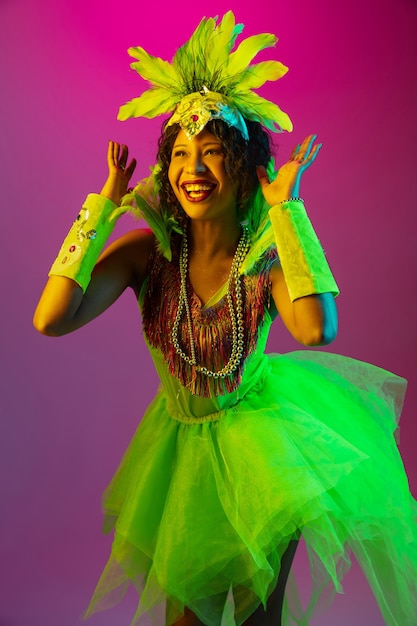 Beautiful young woman in carnival, stylish masquerade costume with feathers dancing on gradient wall in neon