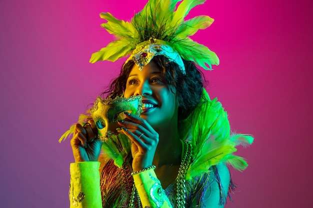 Beautiful young woman in carnival, stylish masquerade costume with feathers dancing on gradient wall in neon