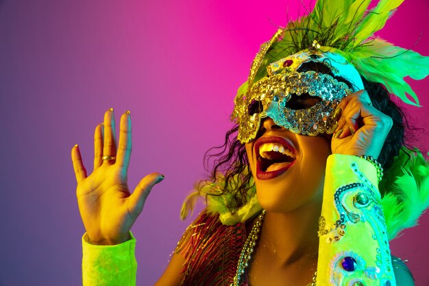 Beautiful young woman in carnival, stylish masquerade costume with feathers on gradient wall in neon light