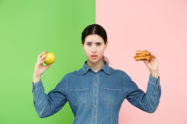 Beautiful young woman choosing between fruits and unhelathy fast food at studio.