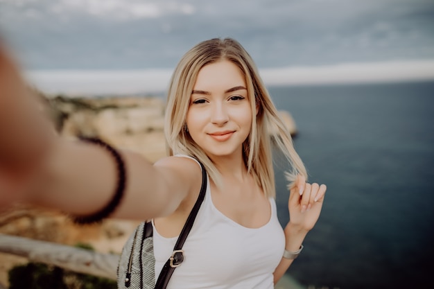 Beautiful young woman doing selfie on the cliff ocean beach