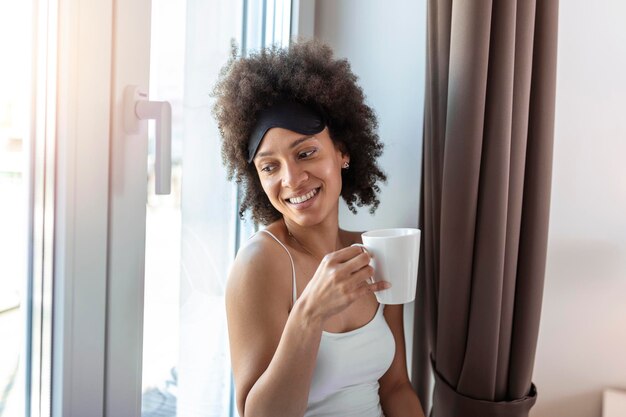 Beautiful young woman drinking coffee and looking through window while sitting at windowsill at home having a perfect cozy morning