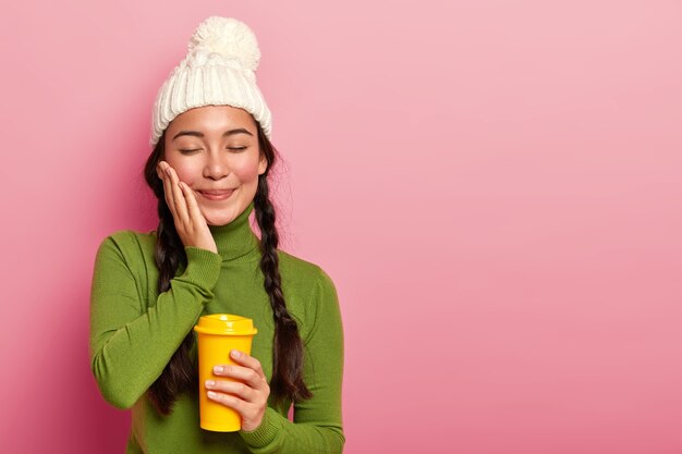 美しい若い女性は使い捨てカップから芳香飲料を飲み、ルージュの頬に触れ、穏やかな外観を持ち、暖かい服を着て、ピンクの背景で隔離