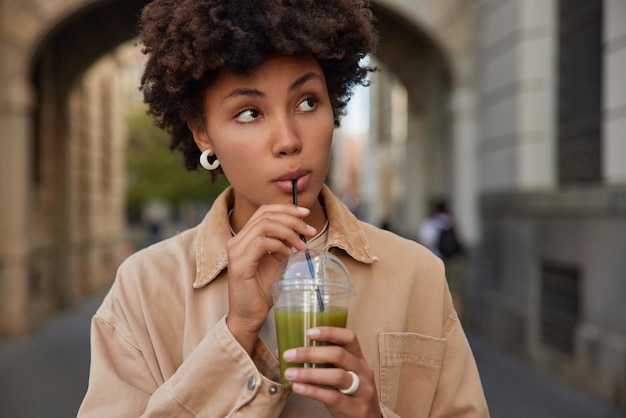 아름다운 젊은 여성은 야외에서 흐릿한 배경을 배경으로 베이지색 재킷 포즈를 취한 거리에 집중된 플라스틱 빨대를 사용하여 유기농 녹색 스무디를 마십니다. 건강 음료와 사람